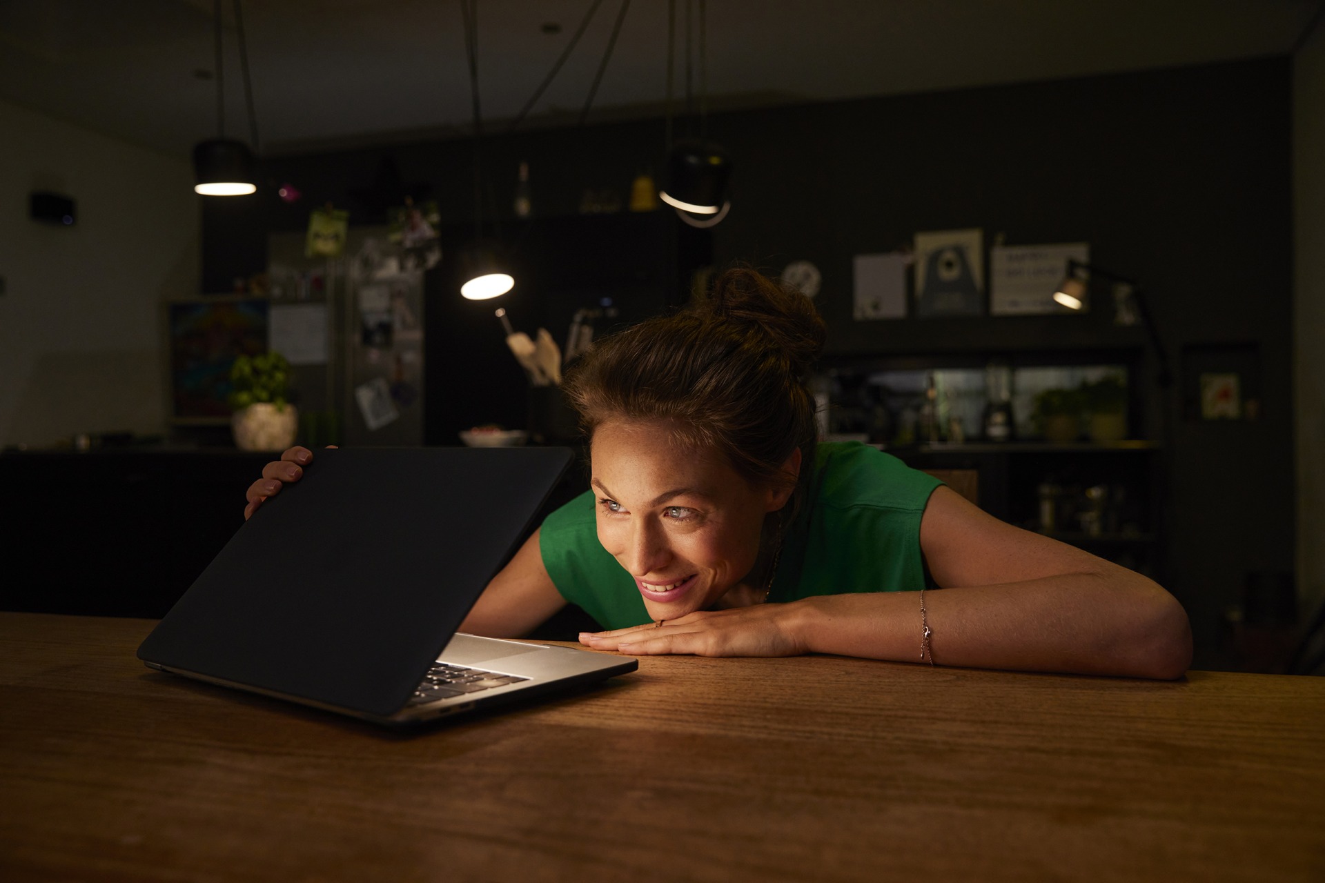 junge Frau sitzt nachts in der Küche und schaut neugierig in leuchtenden Laptop, Lifestylefoto